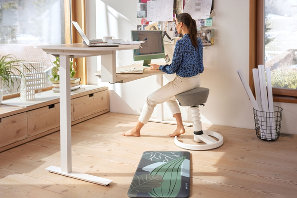 Frau arbeitet an einem höhenverstellbaren Schreibtisch und sitzt dabei auf einem beweglichen ergonomischen Hocker.